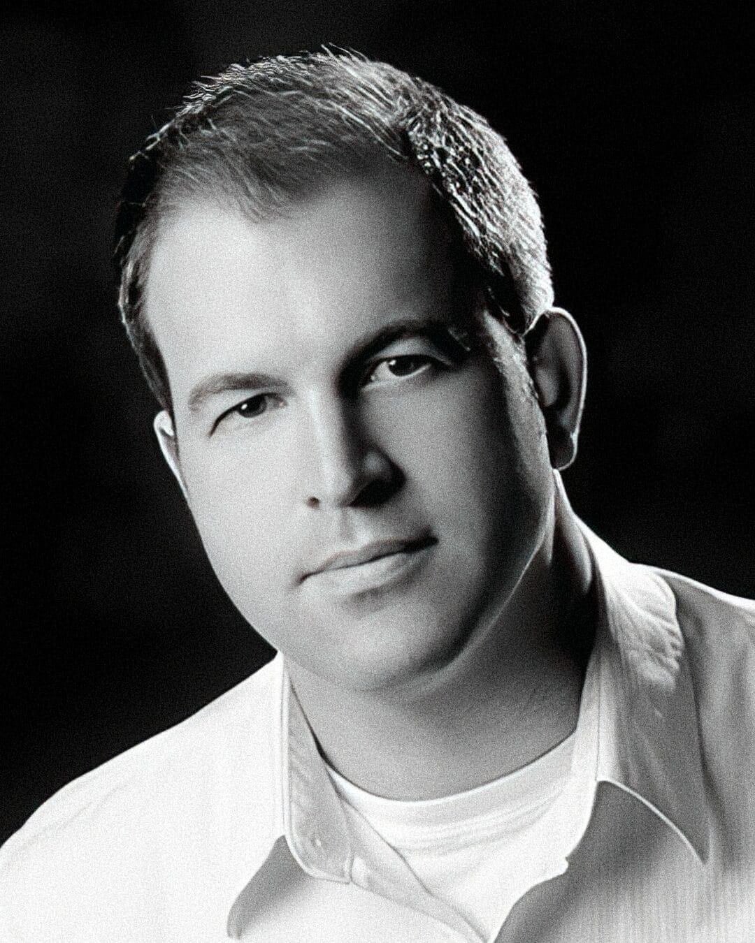 Black and white portrait of a man with short hair, wearing a light-colored collared shirt. He has a neutral expression and is looking directly at the camera. The background is dark, contrasting with his light clothing and complexion.