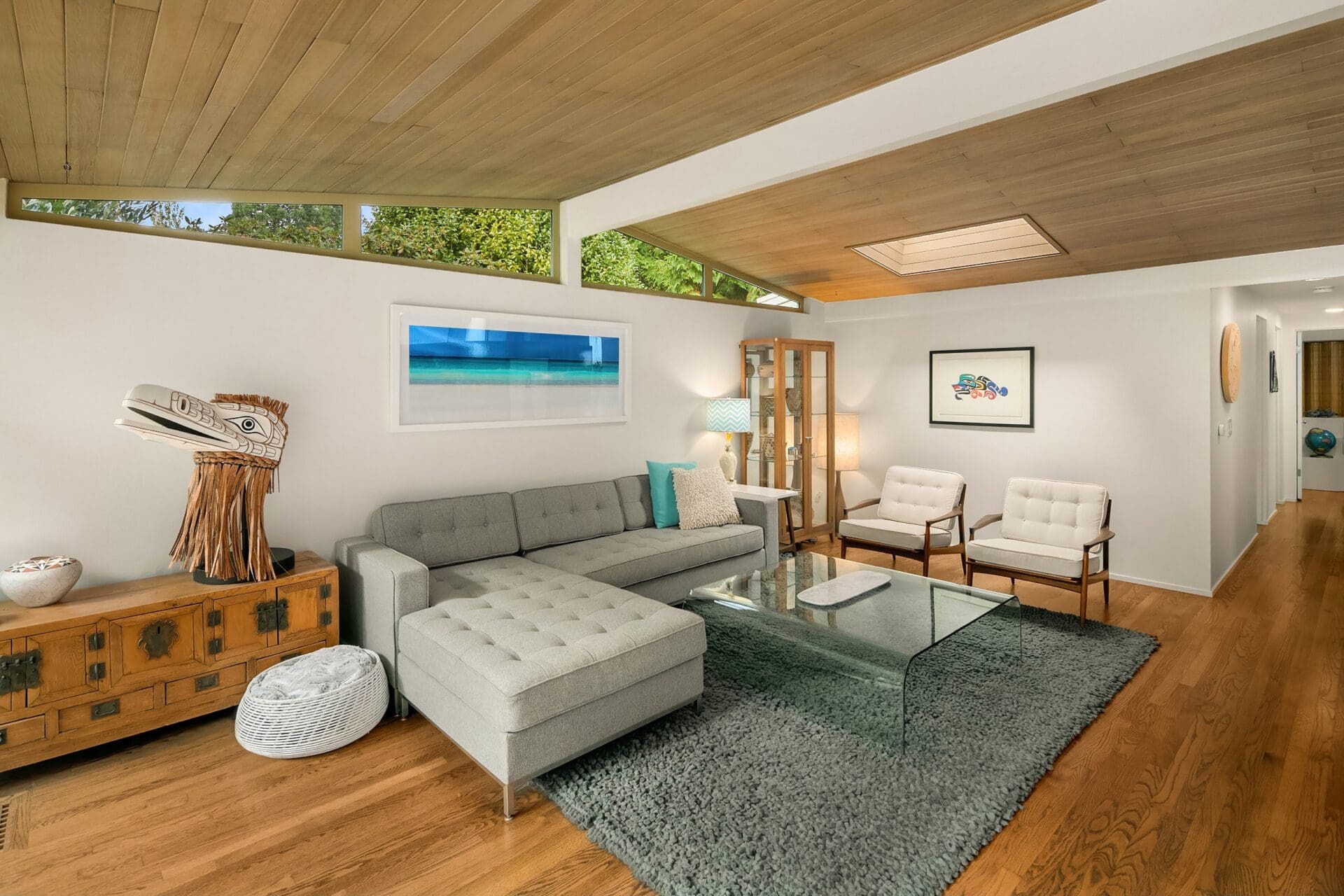 A modern living room with a gray sectional sofa, glass coffee table, and two white armchairs on a gray rug. Wooden ceiling, hardwood floors, and large windows enhance natural light. Decor includes a wall painting, wooden chest, artwork, and a cat bed.