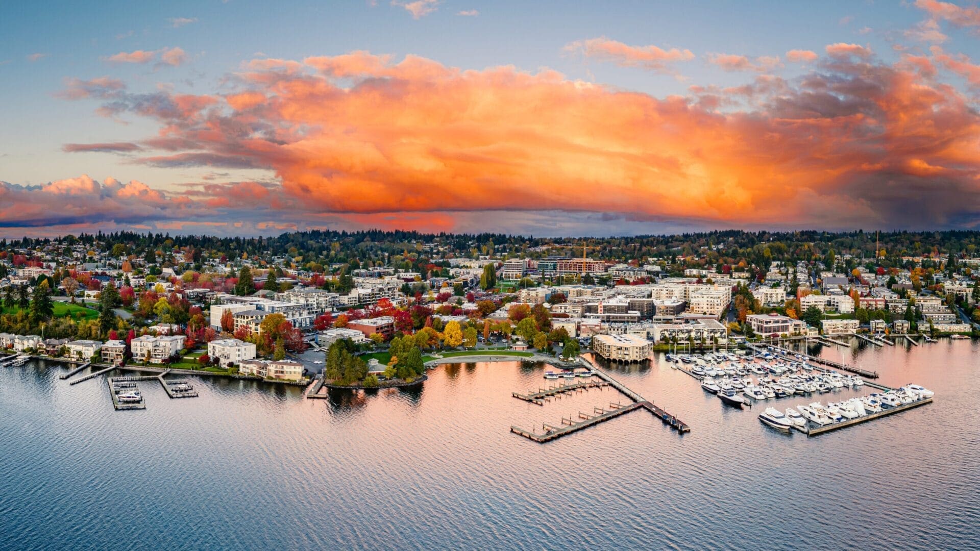 Aerial view of a coastal town featuring a marina filled with boats, surrounded by a calm body of water. The town has numerous buildings, greenery, and autumn-colored trees. A vibrant, orange-hued cloud formation dominates the sky at sunset.