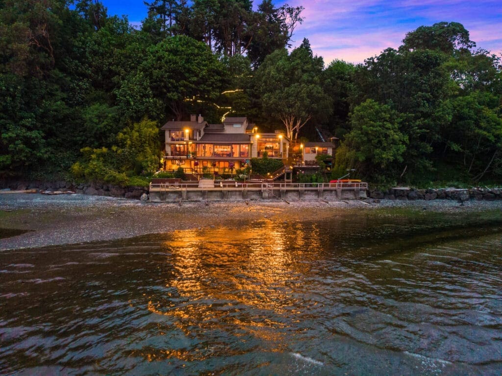 A charming house with lights on, nestled among lush trees and facing a serene body of water at dusk. The warm glow from the lighting is reflected on the calm water, creating a tranquil and inviting scene.