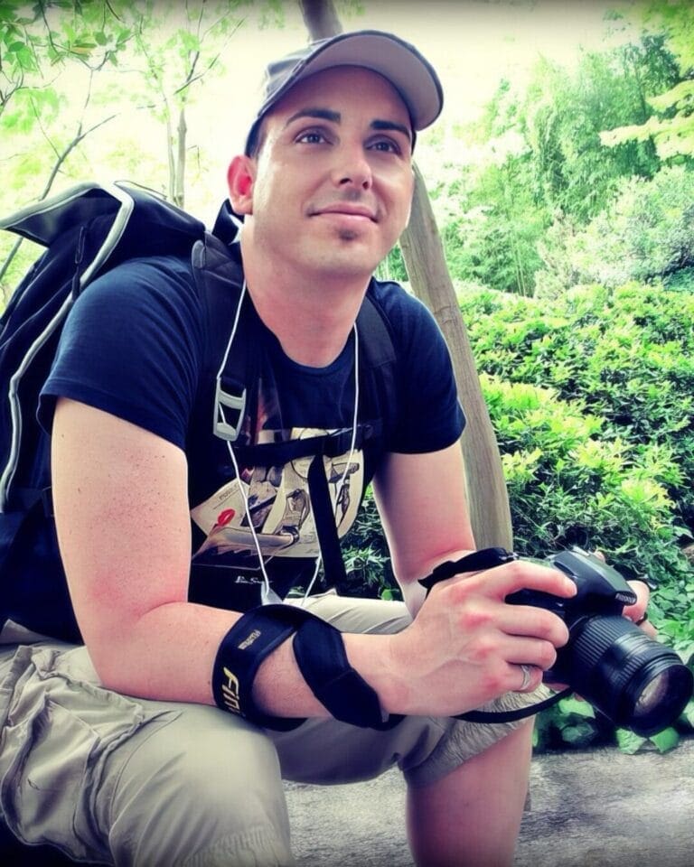 A person wearing a baseball cap, black t-shirt, and beige shorts is sitting outdoors holding a DSLR camera. They are carrying a backpack and have earphones on. Green foliage and trees are in the background, indicating a natural setting.