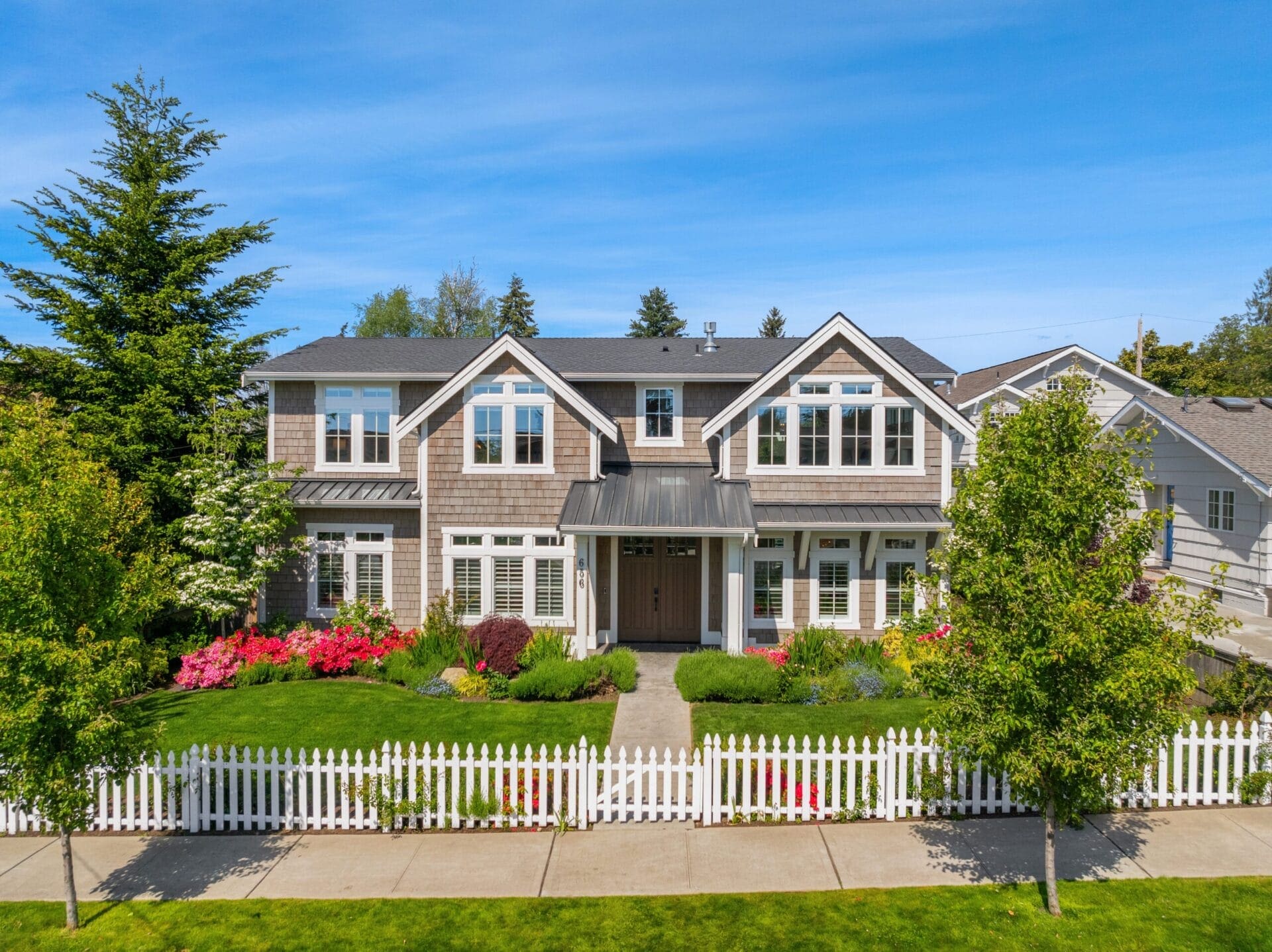 A two-story suburban house with grey shingle siding, white trim, and large windows. It has a manicured front yard with vibrant flowers, green bushes, and trees. A white picket fence surrounds the yard, and a pathway leads to the front door. The sky is clear and blue.