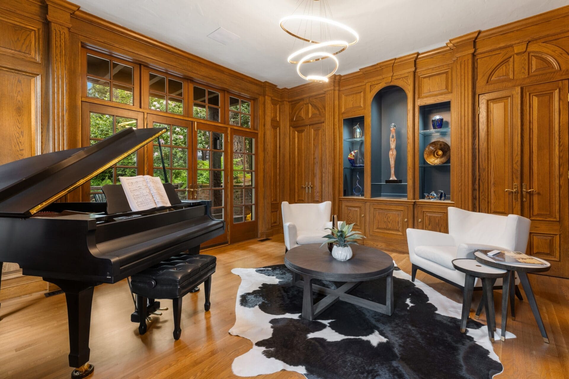 A luxurious room with wooden paneling features a grand piano with sheet music, two white armchairs with a small table between them, and a central round coffee table on a cowhide rug. The built-in wooden shelves display various decorative items.