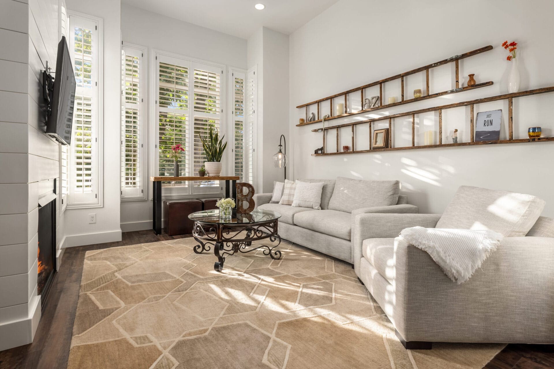 A modern, sunlit living room features a light grey sofa and matching armchair, a glass coffee table, and a large geometric rug. A TV is mounted above a fireplace on the left, while a wooden shelf with various decor items hangs on the right wall. Large windows with white shutters adorn the back wall, letting in ample natural light.