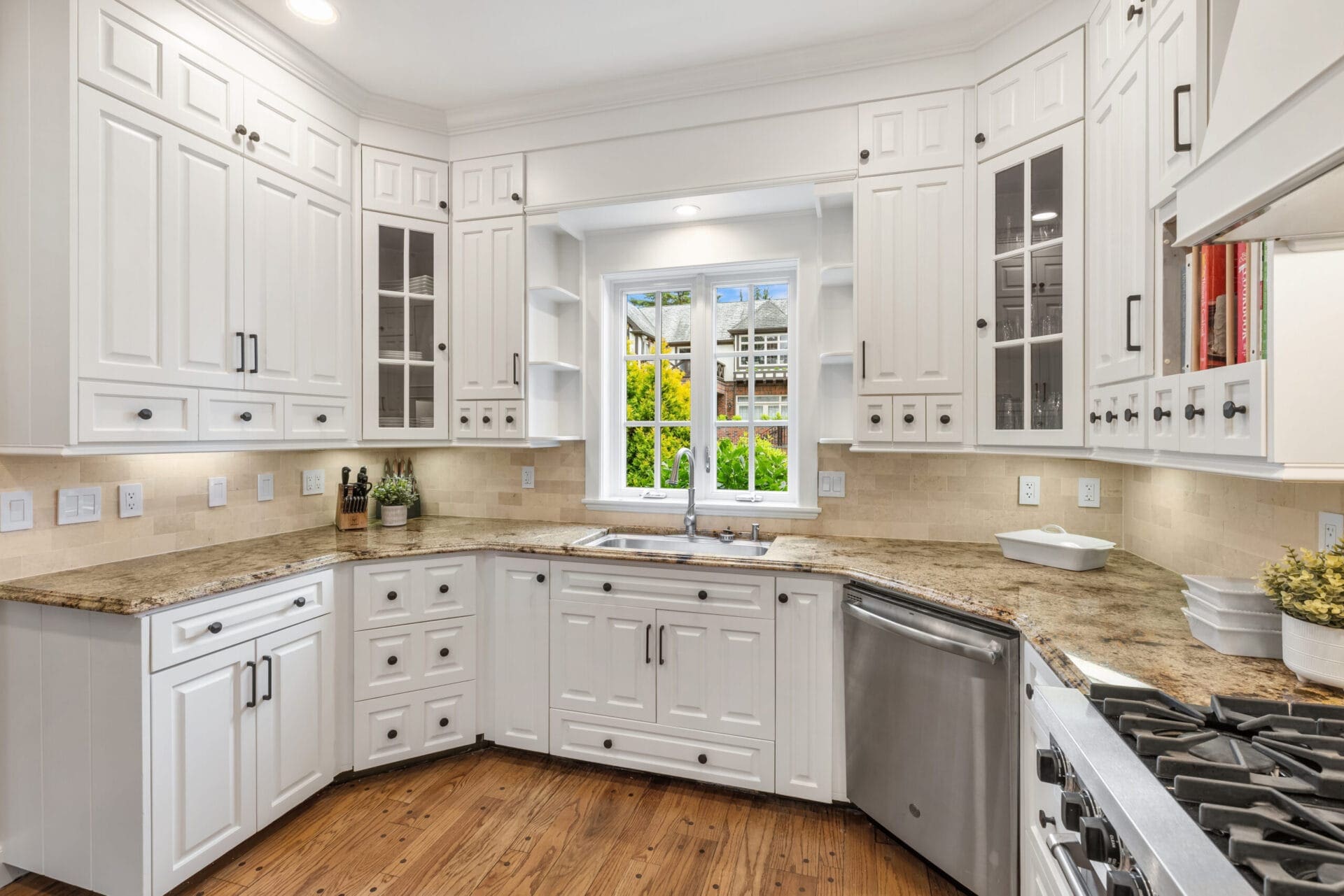 A bright, modern kitchen with white cabinets and a polished beige granite countertop. The kitchen features a large farmhouse sink under a window, stainless steel appliances, and wood flooring. Glass-front upper cabinets and decorative items add to the elegant design.