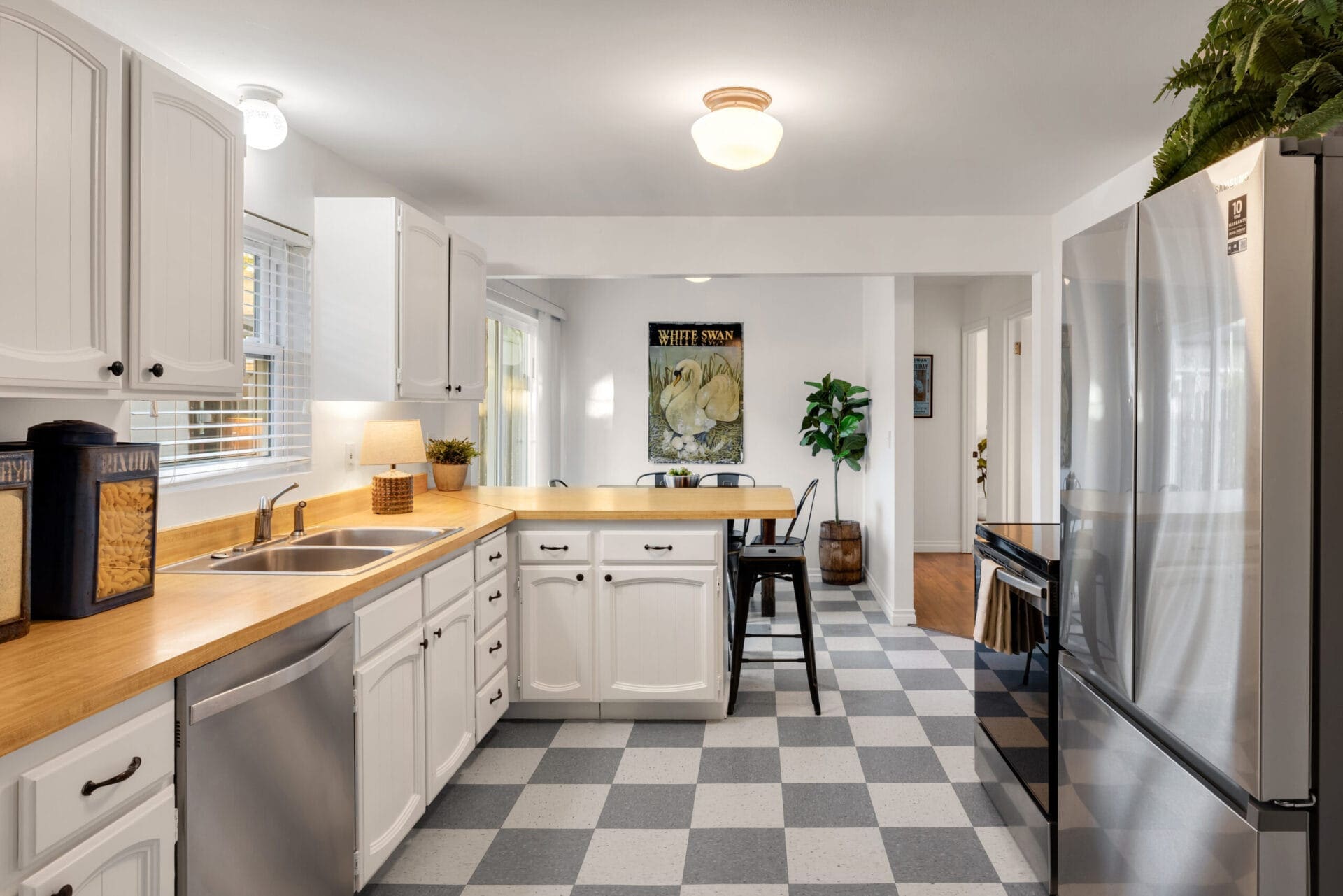 A bright kitchen with white cabinets, wood countertops, and black-and-white checkered flooring. Stainless steel appliances are on the right, and a double-basin sink is on the left. A bar counter with stools, a potted plant, and wall art are in the background.