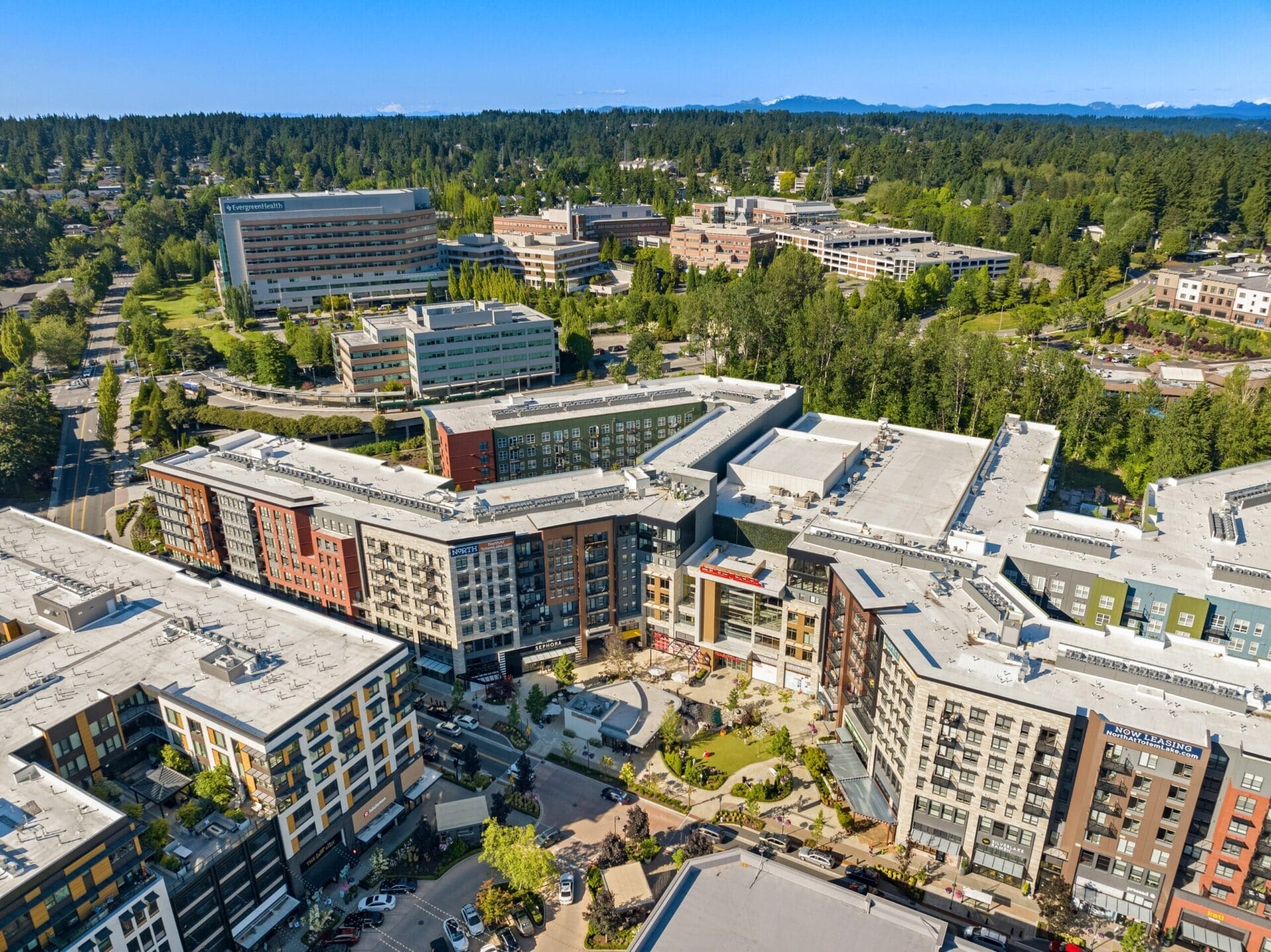 An aerial view of a modern urban area with several large, multi-story buildings arranged around a central courtyard. Trees and greenery are interspersed throughout the development. In the background, a densely forested area extends towards distant mountains under a clear blue sky.