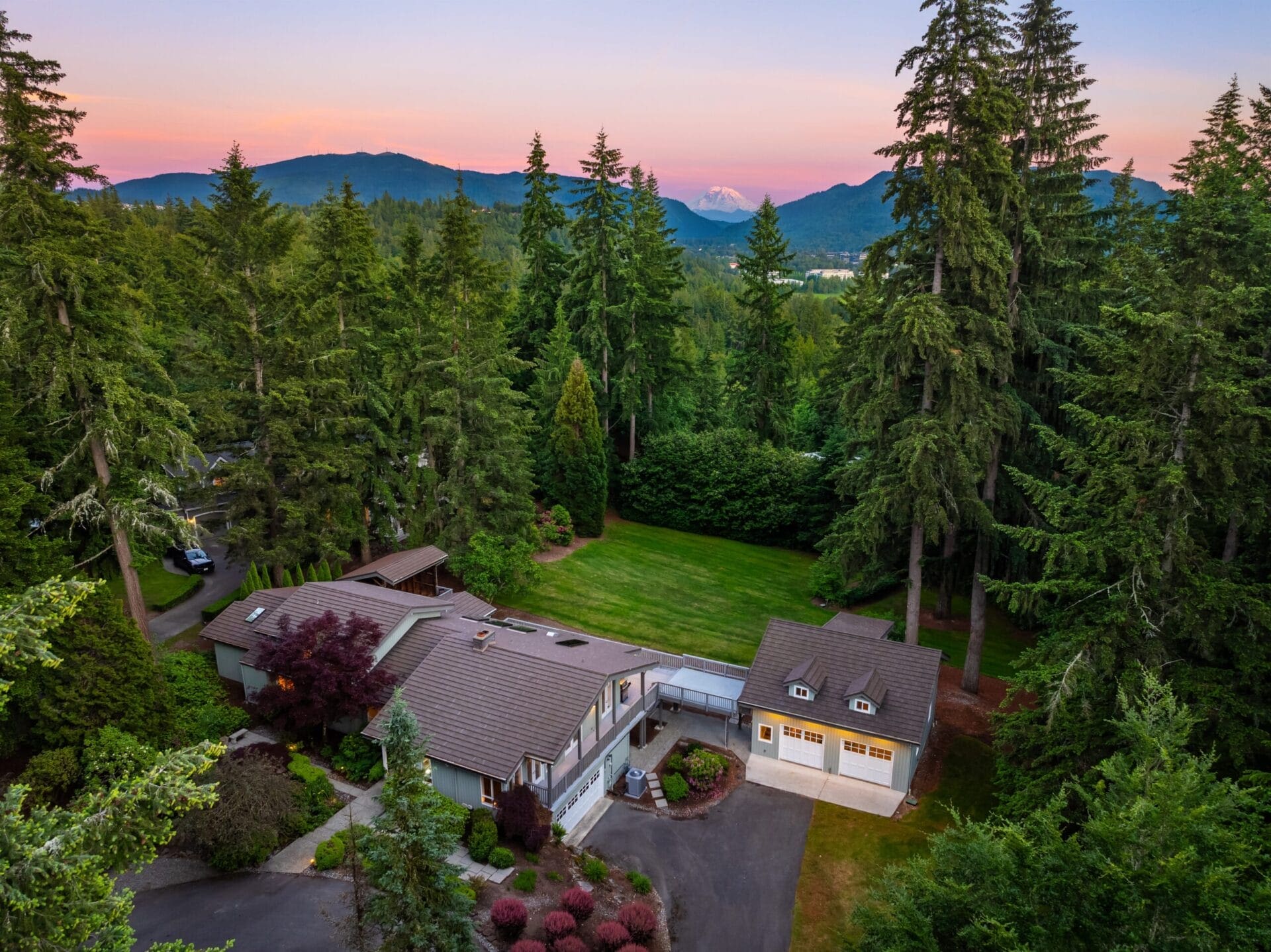 Aerial view of a large house surrounded by dense, tall trees with a well-maintained lawn. The home has a mix of lighter roof sections and includes a driveway leading to the main entrance. In the background, rolling hills and a colorful sunset sky are visible.