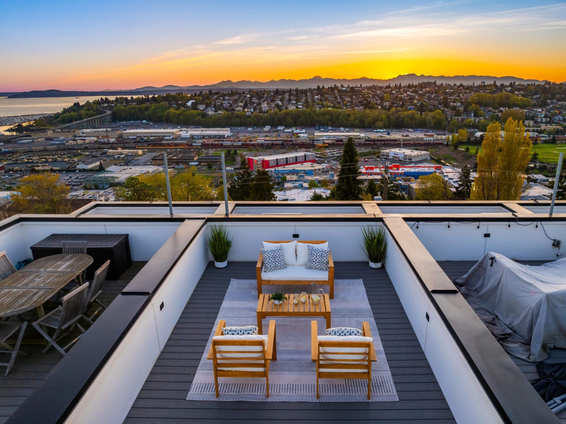 A modern rooftop patio with wooden furniture, potted plants, and a stunning sunset view. The patio features a central seating area with a cushioned sofa, two chairs, and a coffee table. The background shows a cityscape, hilly landscape, and distant mountains.