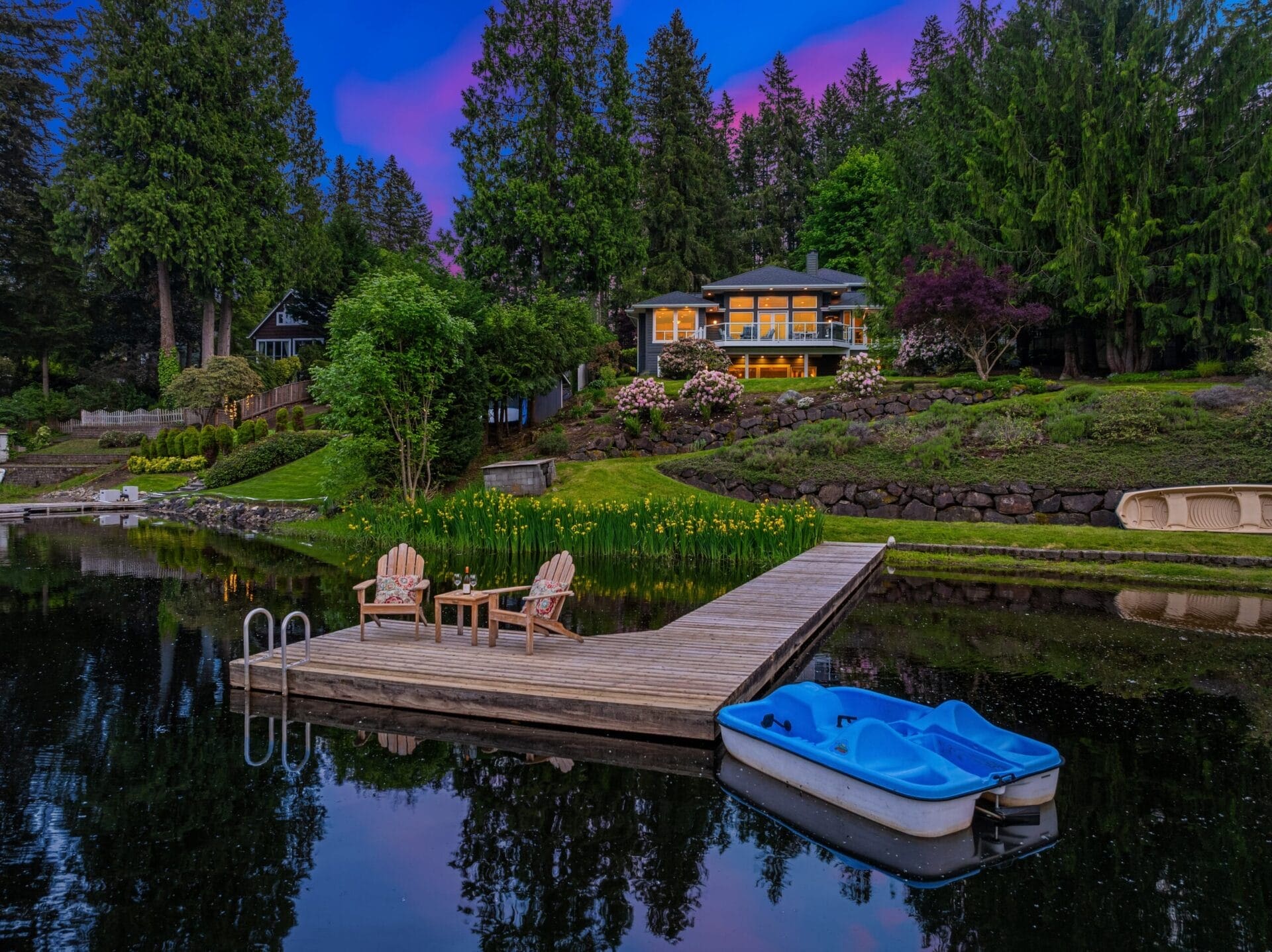 A serene lakeside scene at dusk with a wooden dock extending into calm water. Two Adirondack chairs and a small table are on the dock. A blue paddleboat is tied to the dock. In the background, a well-lit house is surrounded by lush green trees and a vibrant landscape.