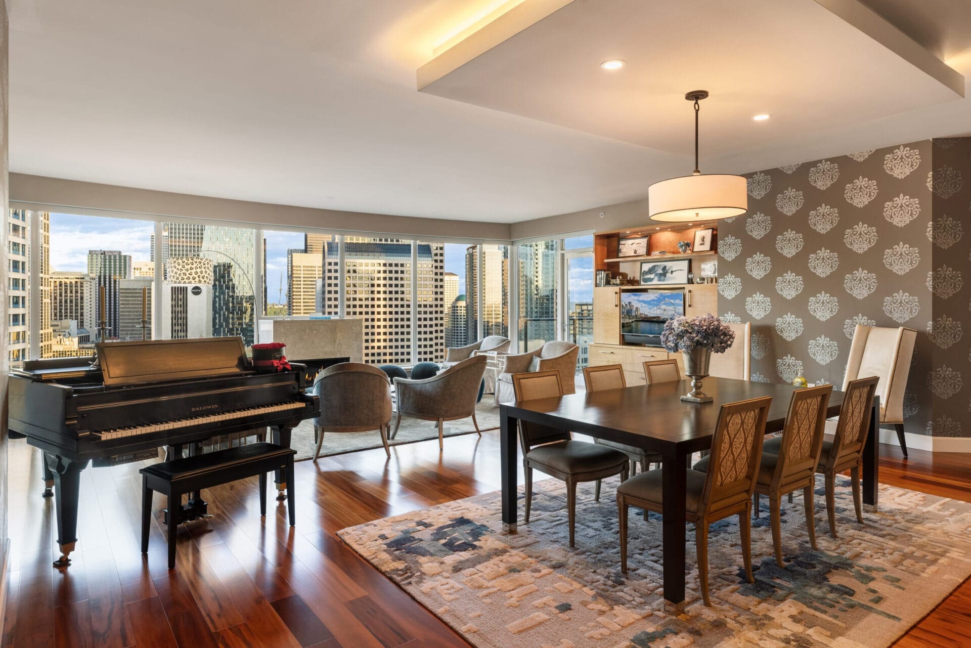 A modern living and dining area with a grand piano on the left, a dining table with six chairs in the center, and a seating area with four armchairs by large windows showcasing a cityscape. The room features patterned wallpaper, hardwood floors, and elegant decor.