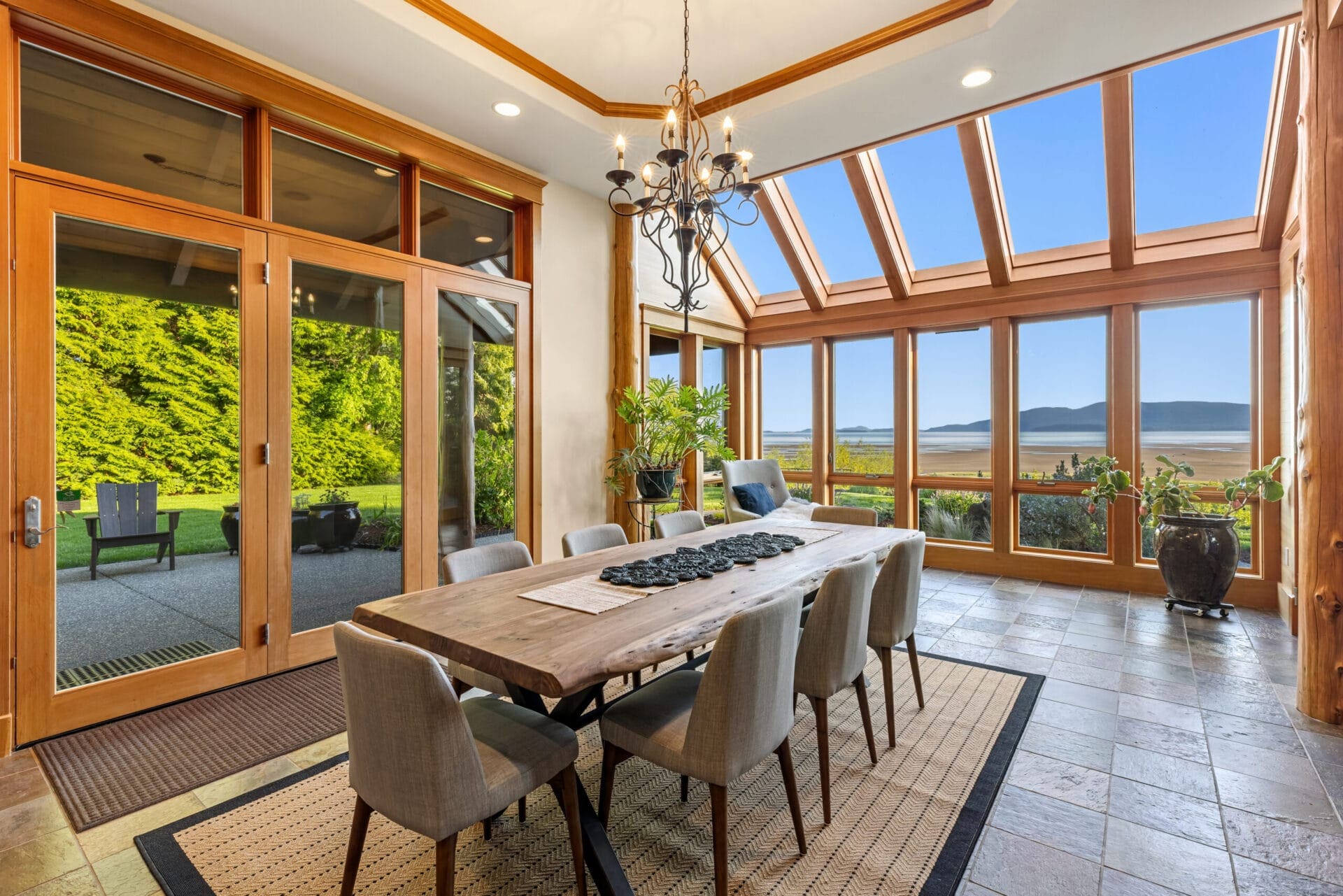 A modern dining room featuring a large wooden table with eight gray upholstered chairs. The room has floor-to-ceiling windows and skylights, offering panoramic views of lush greenery and distant mountains. A dark chandelier hangs above the table.