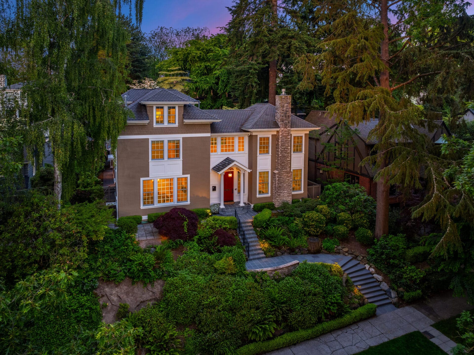 A large, elegant house with beige exterior, white trim, and a stone chimney is surrounded by lush greenery and tall trees. The front yard features a well-maintained garden with shrubs, a walkway leading to the front door, and a set of steps. The windows are lit warmly.