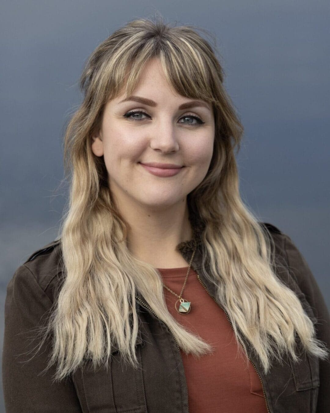 A person with long, wavy blonde hair wearing a brown jacket and an orange top smiles at the camera. The background is an outdoor setting with a muted, out-of-focus view.