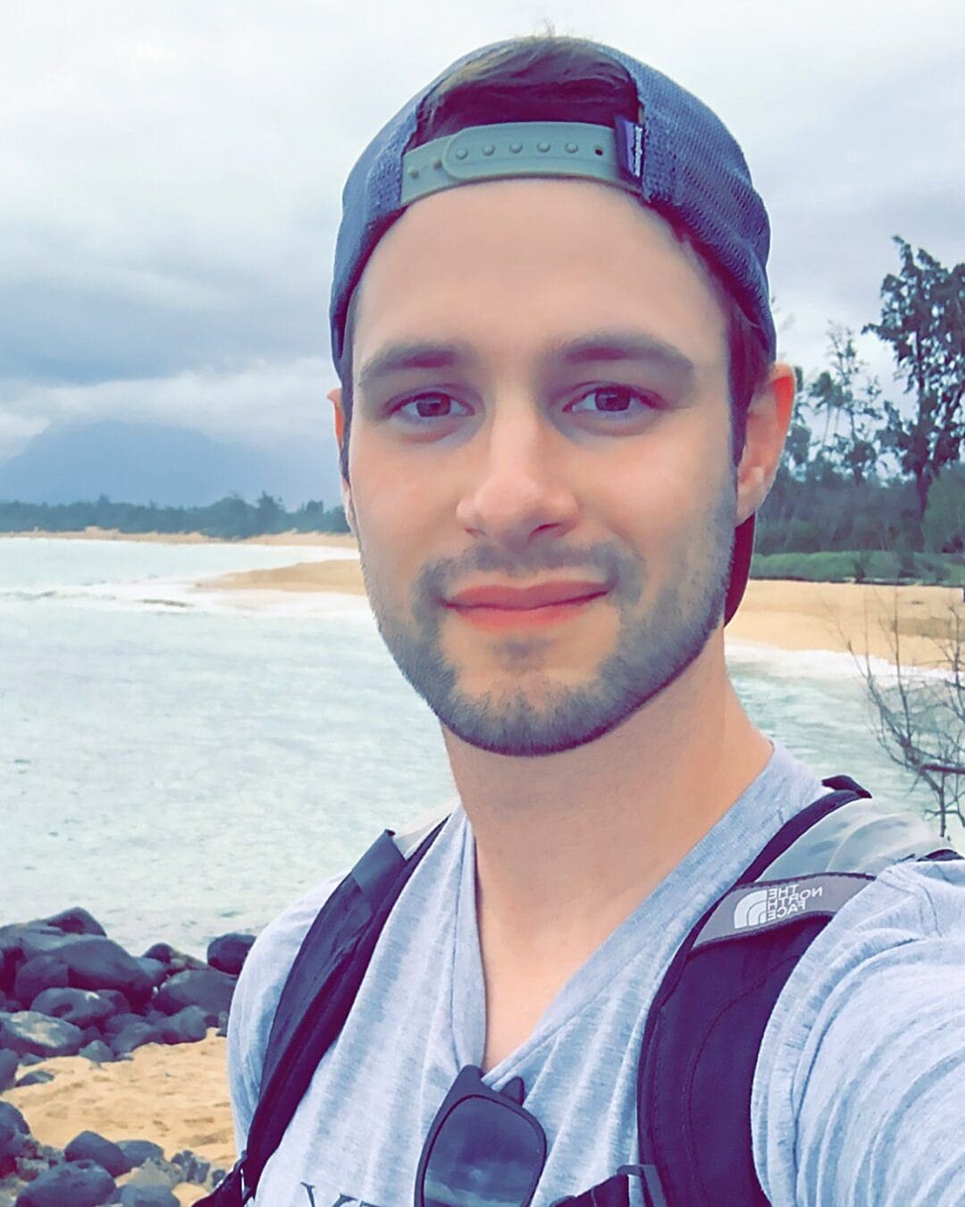 A young man with a short beard and wearing a gray shirt, sunglasses hanging from his shirt collar, a backward gray cap, and a black backpack, smiles at the camera. He stands on a rocky beach with overcast skies and the ocean in the background.