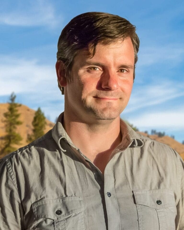 A man with short brown hair stands outdoors in front of a backdrop of trees and hills. He is wearing a light grey button-up shirt and is smiling slightly while looking at the camera. The sky behind him is blue with some wisps of clouds.