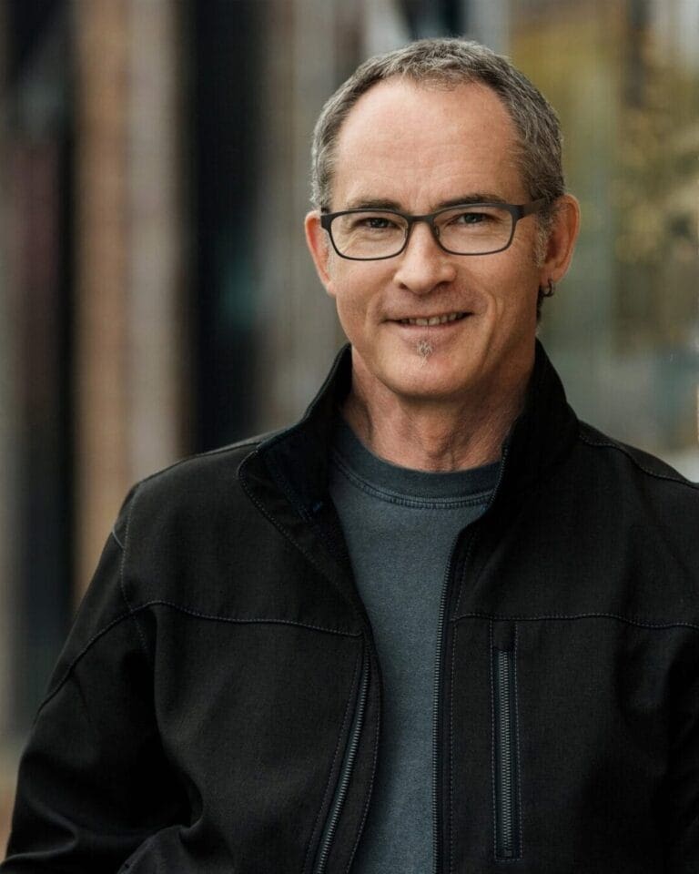 A man with short gray hair, glasses, and a small beard is smiling at the camera. He is wearing a black jacket over a gray shirt and an earring in his left ear. The background is blurred, showing an urban scene.