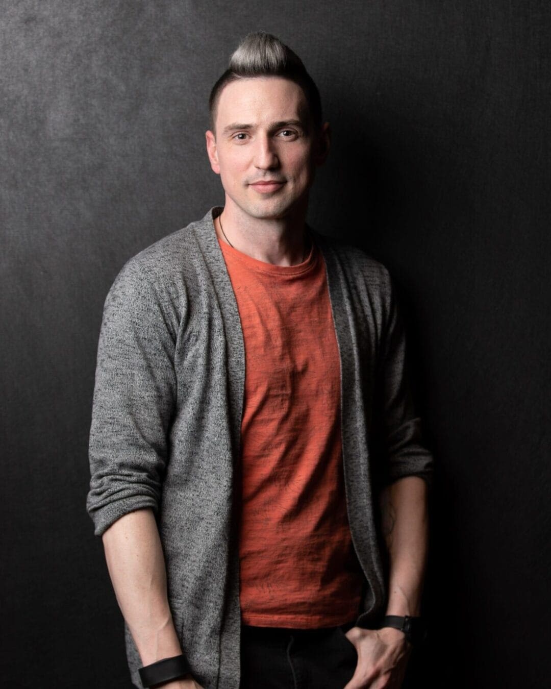 A man with short, styled hair poses against a dark background. He wears a gray cardigan over an orange shirt, and has bracelets on both wrists. He has a relaxed expression with his hands in the pockets of black trousers.