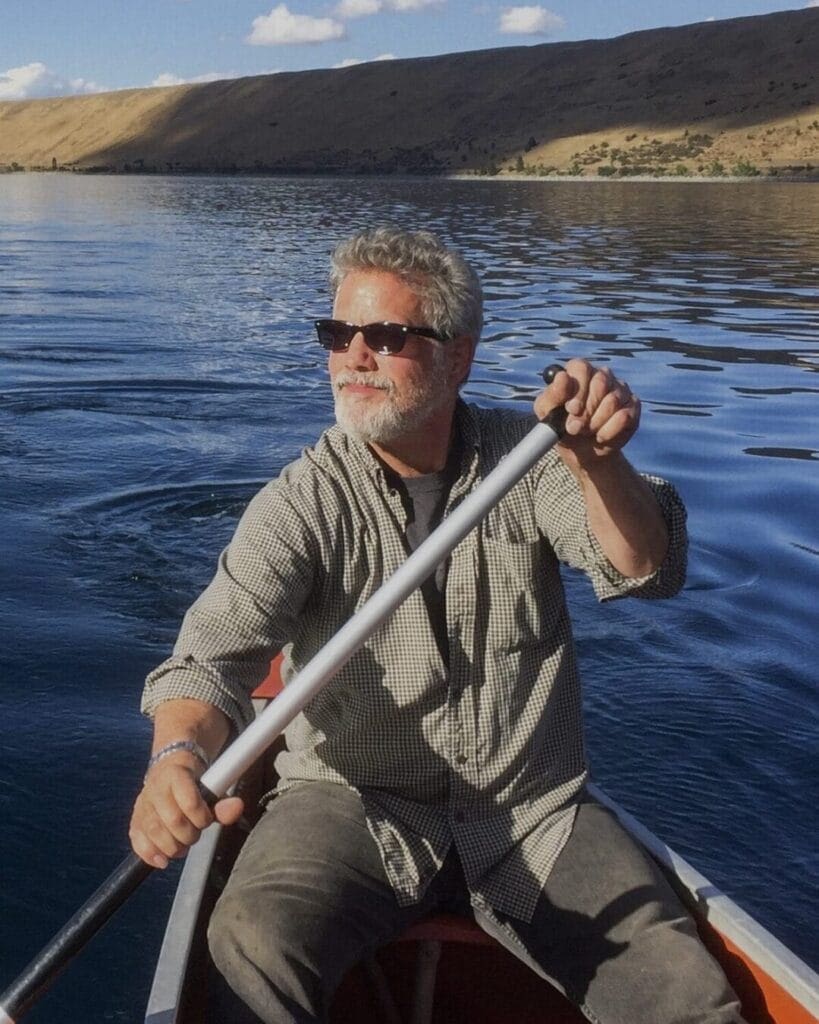 A man with gray hair and a beard, wearing sunglasses and a checkered shirt, smiles as he rows a boat on a calm lake. The background features a hillside with sparse vegetation under a clear blue sky.