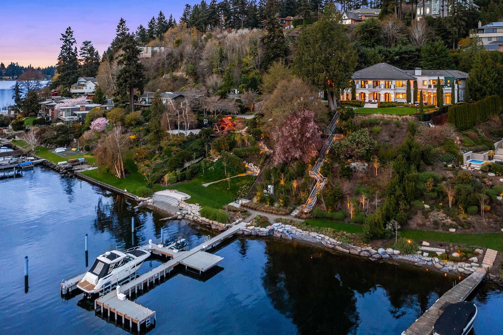 A scenic lakeside property featuring a large, elegant house with illuminated windows. The property is surrounded by lush trees and gardens, with a staircase leading down from the house to a private dock with a boat. Other homes and a forested area are visible in the background.