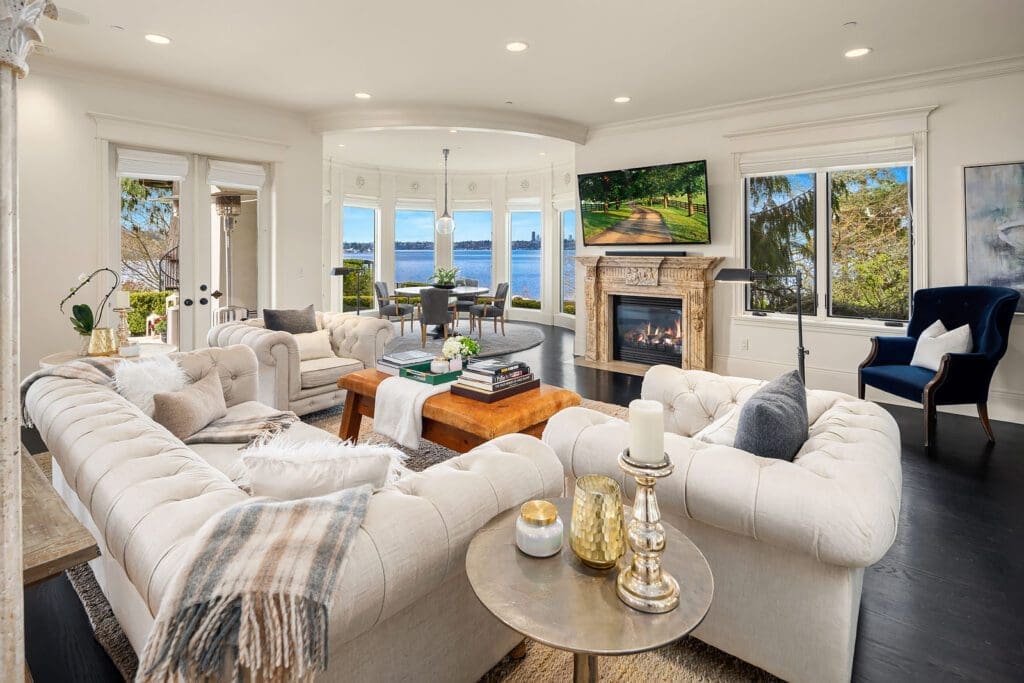 A spacious, elegant living room featuring a large beige sectional sofa adorned with cushions and blankets, a wooden coffee table, a fireplace with a TV mounted above, and expansive windows offering a stunning view of a lake and lush greenery.