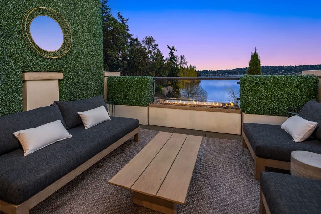 A cozy outdoor seating area with dark gray cushioned sofas and a wooden coffee table, centered around a modern rectangular fire pit. The space is surrounded by green foliage walls, and offers a scenic view of a lake at sunset.