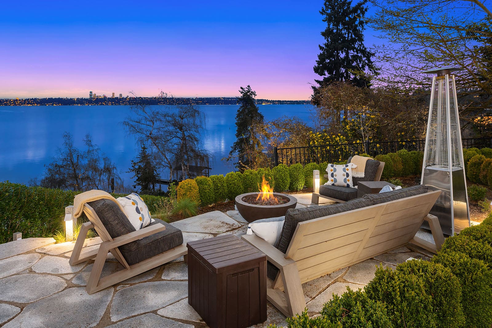 Outdoor patio with comfortable chairs surrounding a fire pit overlooking a serene lake at sunset. The vibrant twilight sky and distant cityscape create a tranquil ambiance. Trees and shrubs border the stone patio, enhancing the natural beauty of the scene. Twilight replacement.