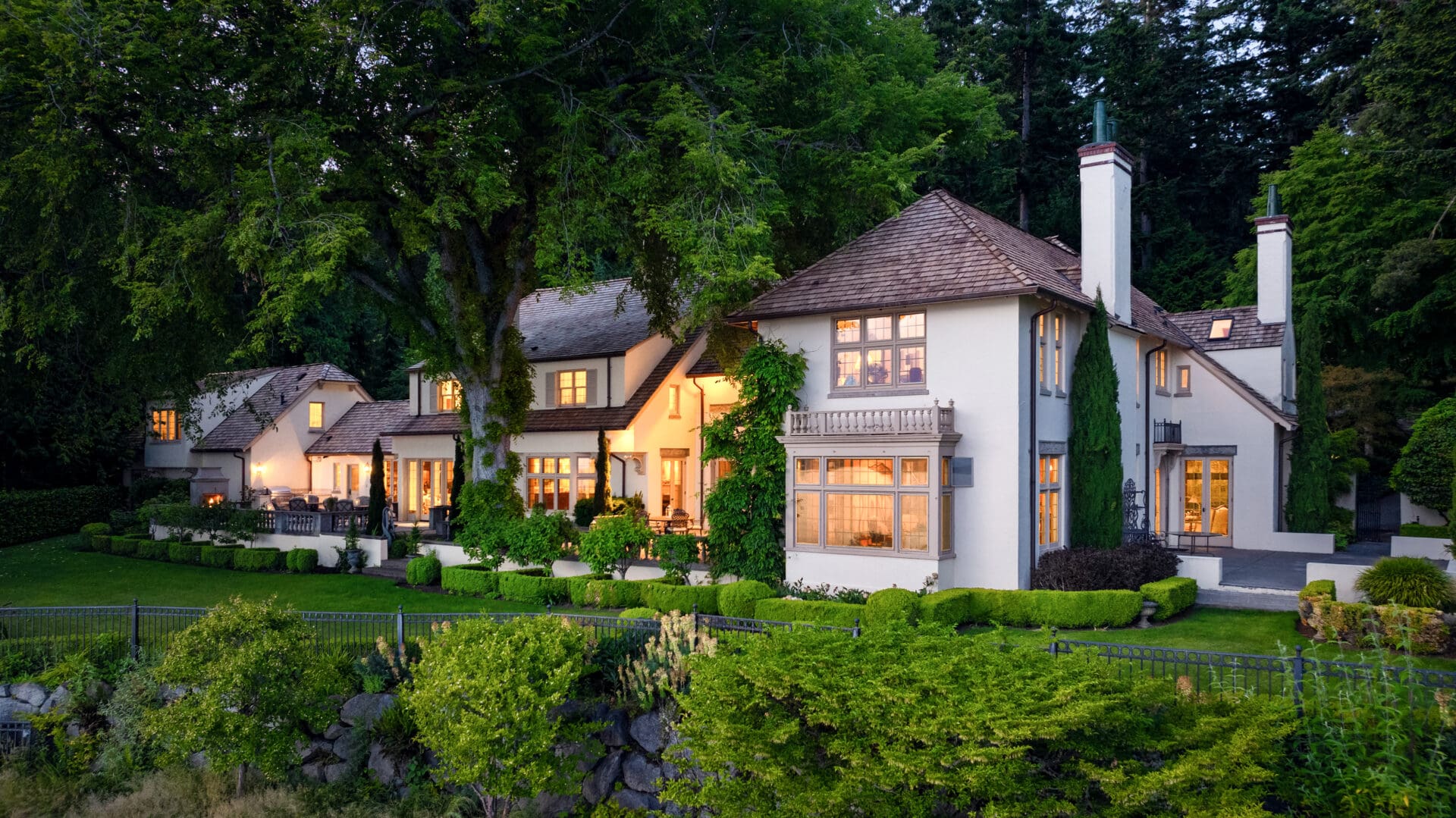 A large, elegant house with a stone exterior and multiple peaked roofs is nestled in a lush, wooded area. The Tim Van Asselt Edwin Ivey estate features numerous windows, balconies, and exterior lighting that gives it a warm and inviting glow. Manicured gardens and a wrought iron fence surround the property.