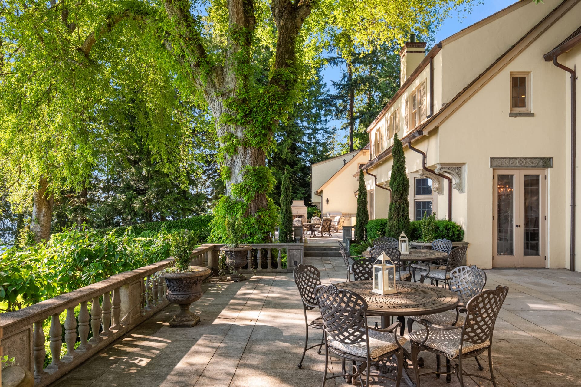 A spacious and elegant outdoor patio of a large house, part of the renowned Tim Van Asselt Edwin Ivey estate, with light-colored walls. The patio features metal tables and chairs, decorative lanterns, and is surrounded by lush greenery and large trees. Stone railings border the patio, offering picturesque views of the garden.