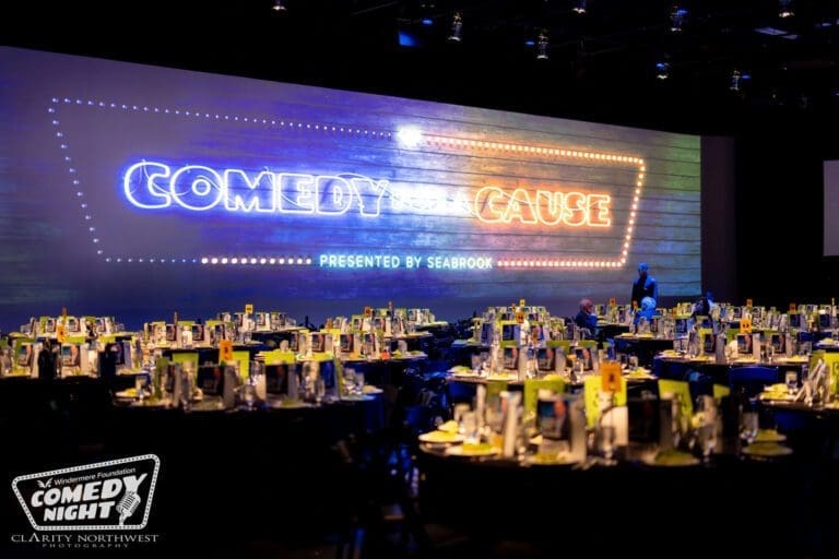 Event space set up for a comedy night, featuring round tables adorned with vibrant green menus and elegant table settings, illuminated by a large neon sign reading "Comedy for a Cause," in a modern venue in the Pacific Northwest.