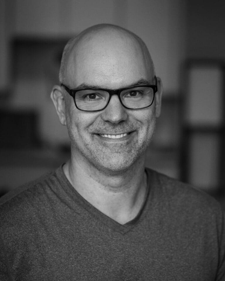 A smiling man in a casual gray shirt, set against a softly blurred interior background, showcasing a modern, minimalist design. Ideal for a real estate profile in Seattle or the Pacific Northwest.
