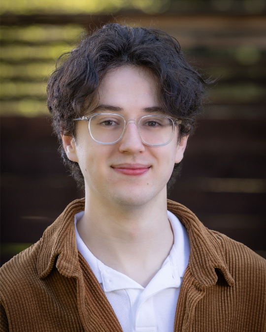 This image features a young man with curly hair and glasses, smiling warmly against a blurred outdoor background, conveying a friendly and approachable vibe typical of the Pacific Northwest.