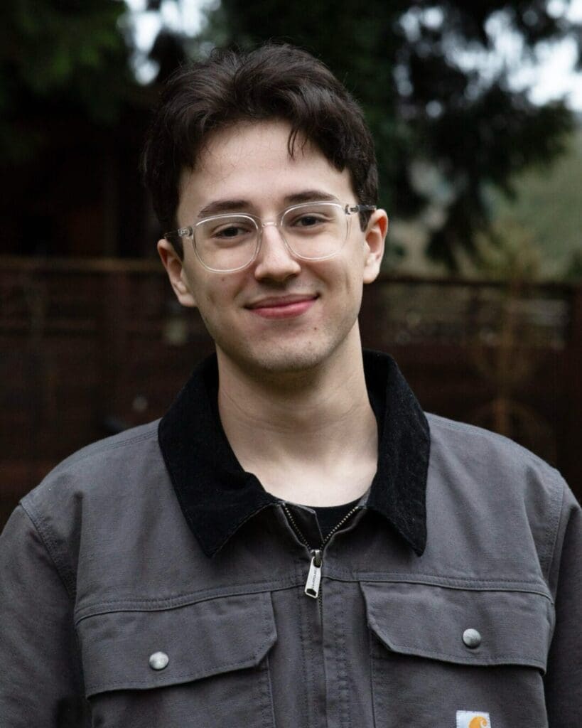 A young man smiling outdoors in a casual gray jacket, surrounded by greenery in the Pacific Northwest.