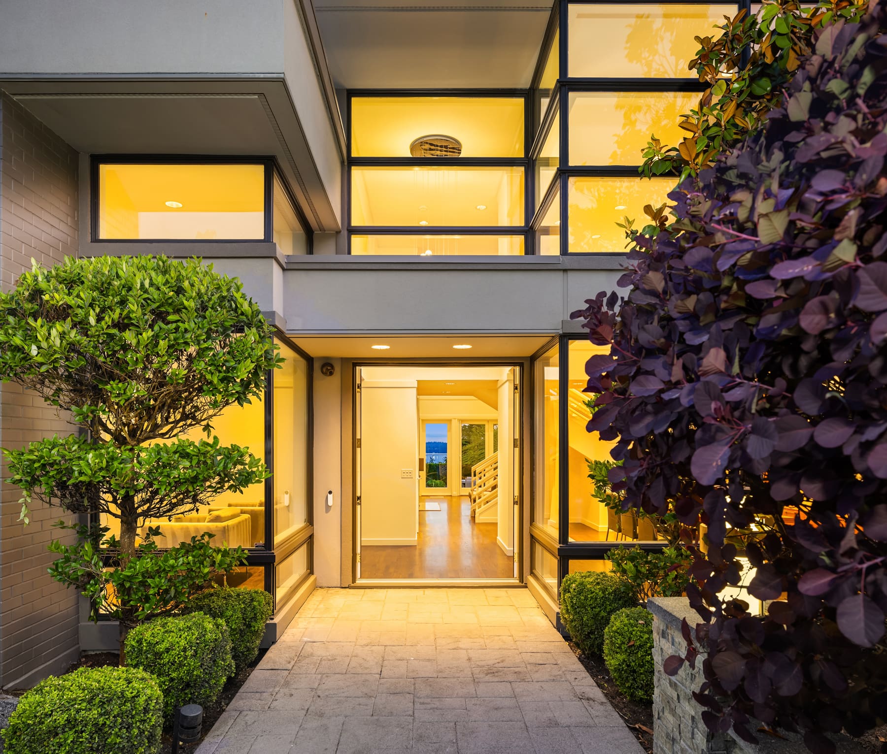 Modern exterior entrance of a contemporary home in the Pacific Northwest, featuring large glass windows, warm lighting, and manicured landscaping with vibrant greenery.