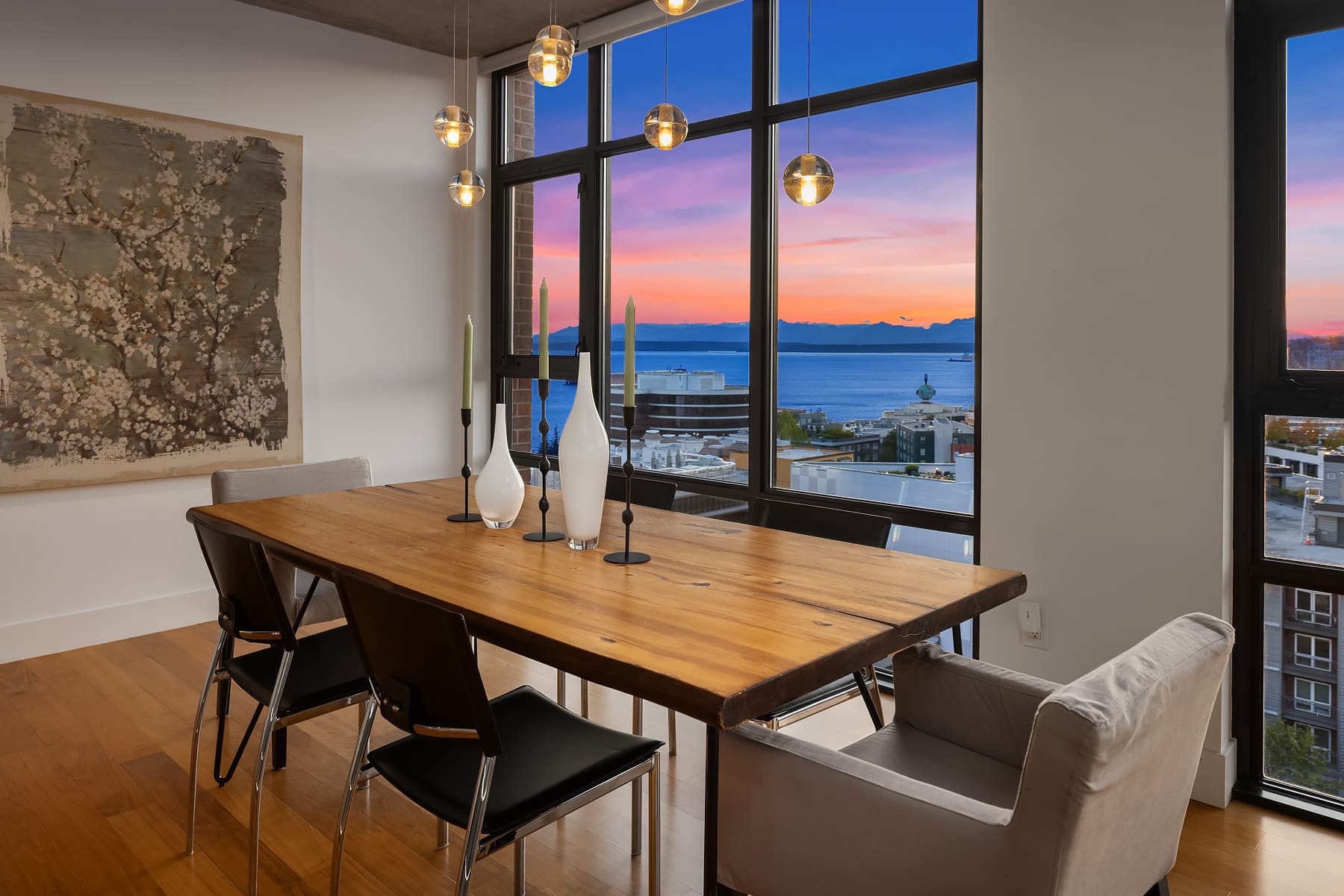 Modern dining room with a wooden table, stylish chairs, and artistic decor, featuring large windows that showcase a stunning sunset view over Seattle's waterfront.