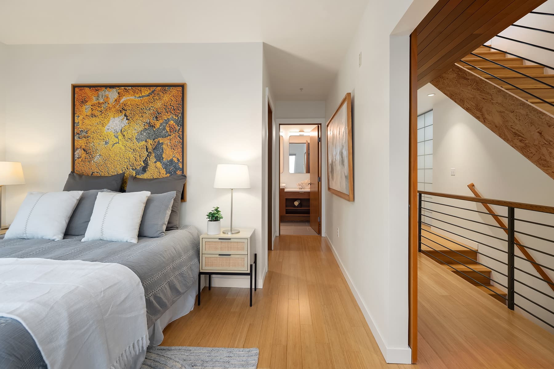 Modern bedroom with bamboo flooring, featuring a stylish bed with gray linens and decorative pillows, complemented by contemporary artwork and soft lighting. The space leads to a hallway and staircase, creating an inviting atmosphere in the Pacific Northwest.