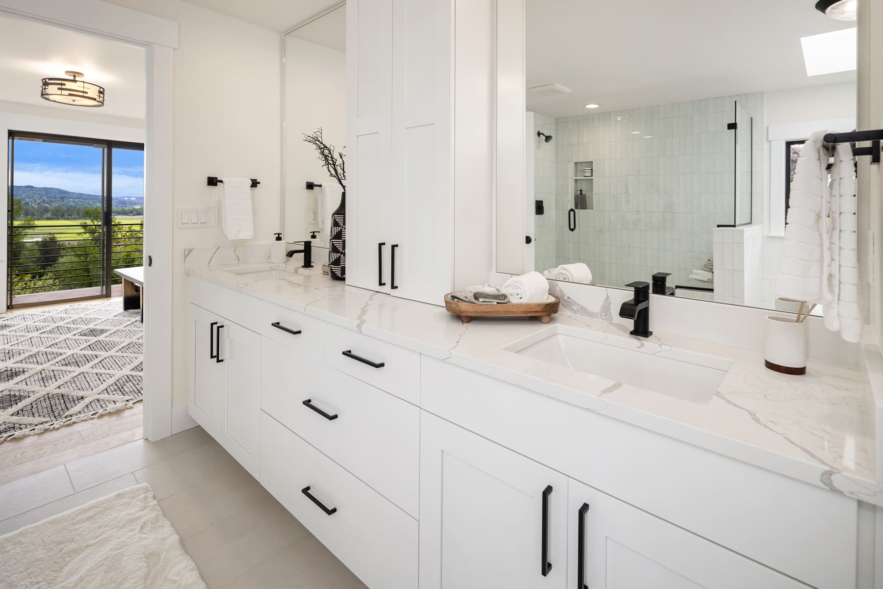 Modern bathroom with sleek white cabinetry, marble countertops, and black fixtures, featuring a large mirror and a view of the Pacific Northwest landscape.