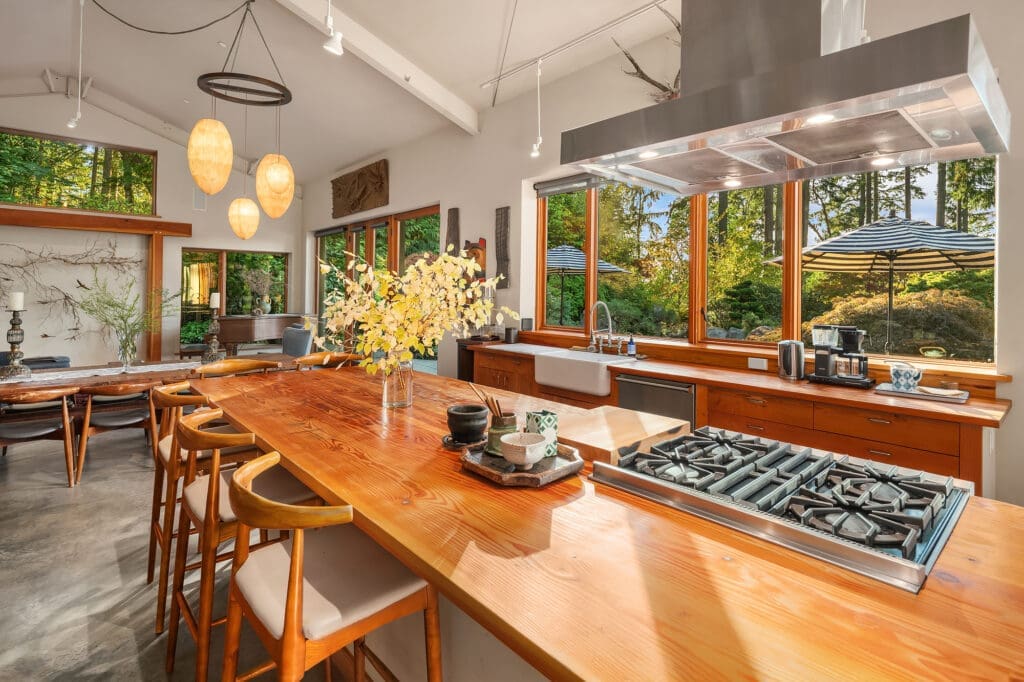 Matt Dillon Vashon Island home photography. Modern kitchen with a spacious wooden island, sleek gas range, and large windows showcasing lush greenery, designed for a warm and inviting atmosphere in the Pacific Northwest.