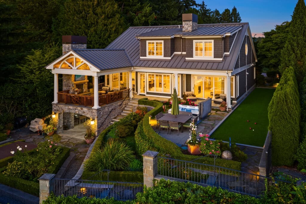 Kell Morrissey's Medina photography. Elegant exterior of a modern home in the Pacific Northwest, featuring stone accents, a spacious patio with outdoor seating, and beautifully landscaped gardens, illuminated at twilight.