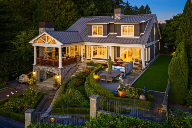 Kell Morrissey's Medina photography. Elegant exterior of a modern home in the Pacific Northwest, featuring stone accents, a spacious patio with outdoor seating, and beautifully landscaped gardens, illuminated at twilight.