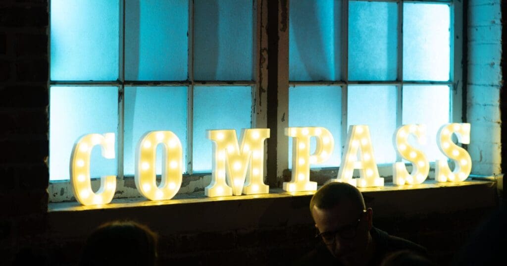 Clarity Northwest Events Team. Illuminated sign spelling "COMPASS" against a backdrop of large, blue-tinted windows in a modern urban space, creating a stylish ambiance, ideal for events in Seattle.