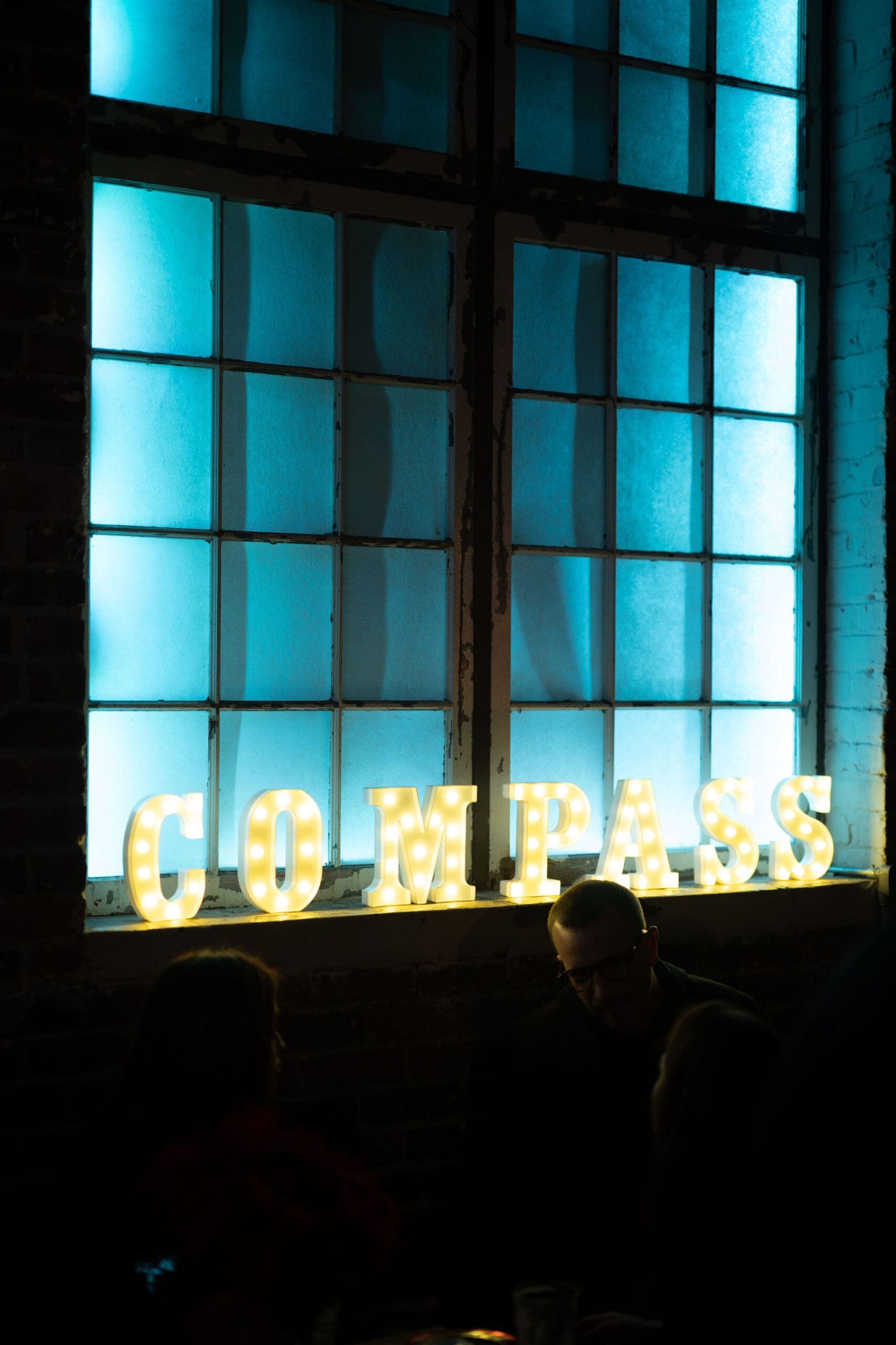 Clarity Northwest Events Team. Illuminated sign spelling "COMPASS" against a backdrop of large, blue-tinted windows in a modern urban space, creating a stylish ambiance, ideal for events in Seattle.