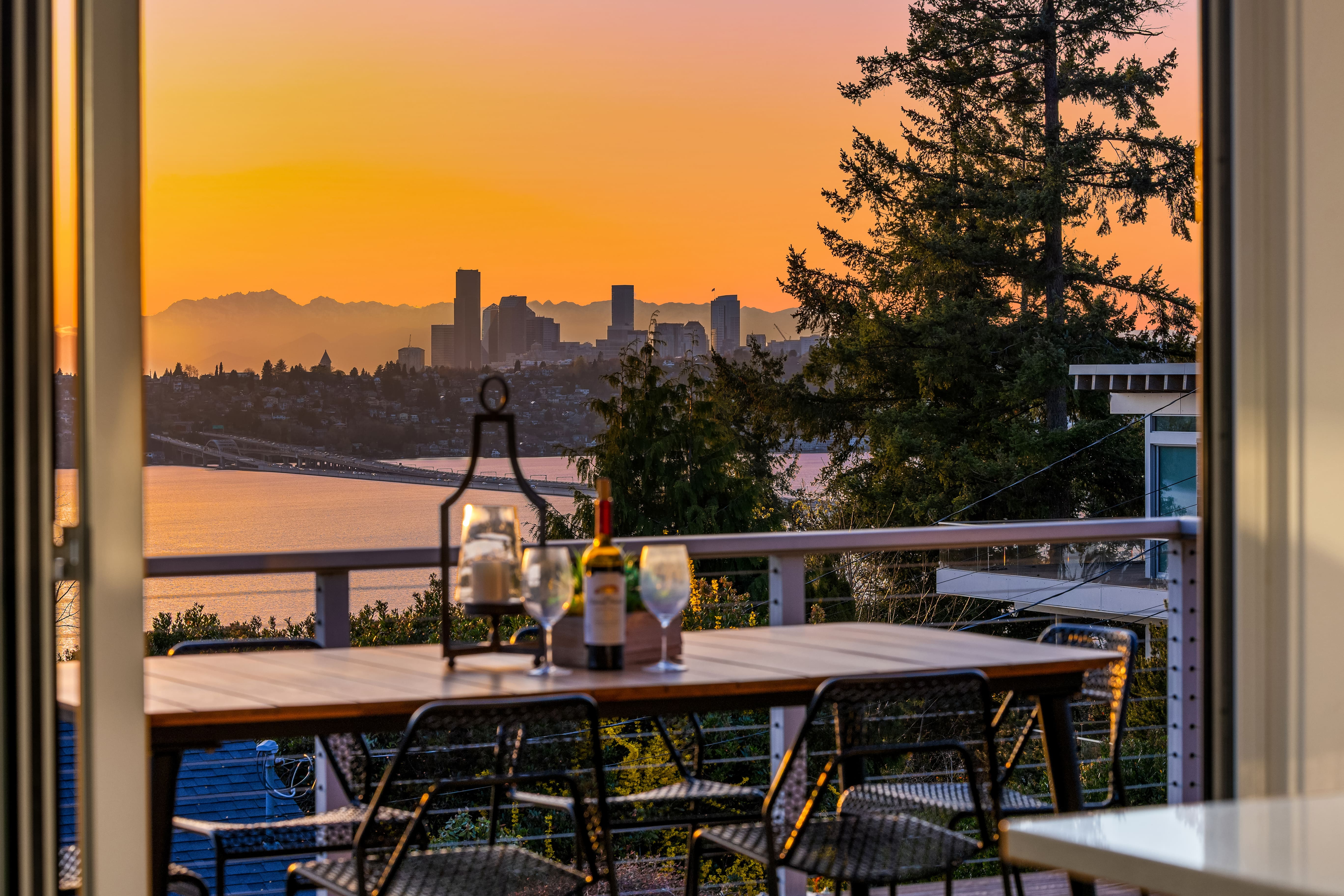 Stunning sunset view from a modern outdoor dining area overlooking Seattle's skyline, featuring a wooden table set with wine and glasses, surrounded by lush greenery.