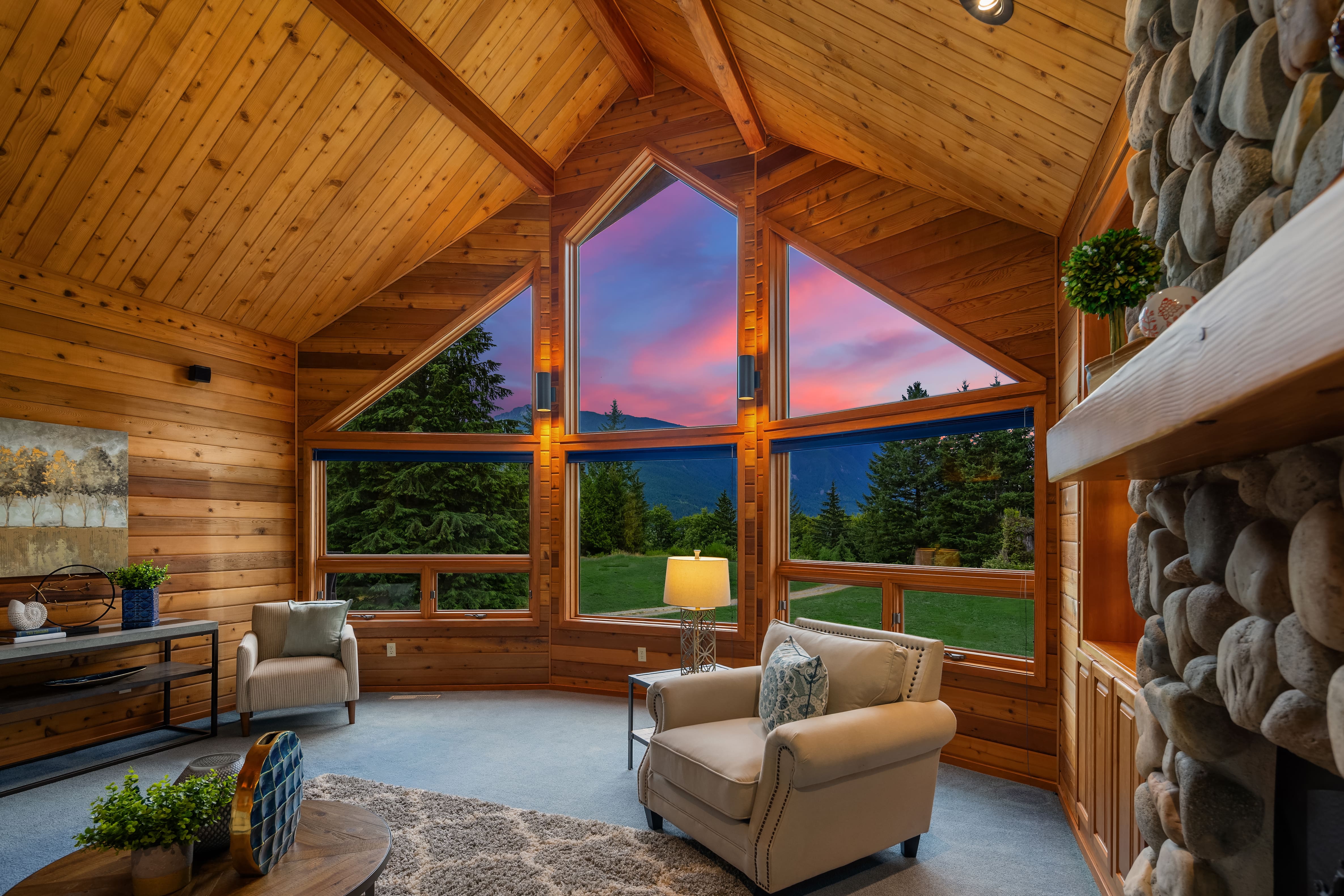 Cozy living room with large triangular windows showcasing a stunning sunset in the Pacific Northwest, featuring warm wood paneling and a stone fireplace for an inviting atmosphere.