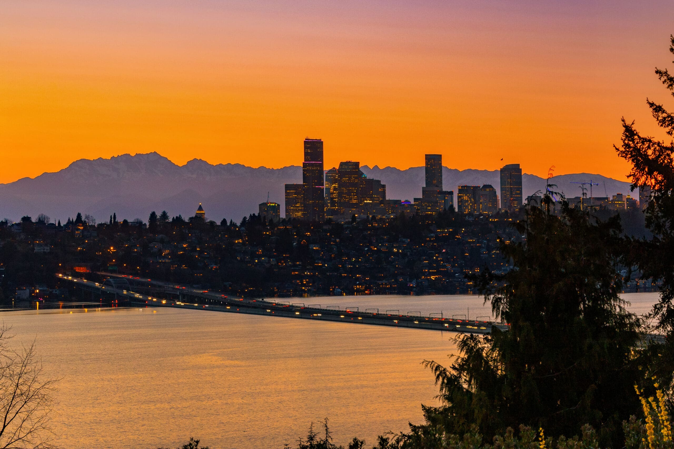 Twilight skyline view of Seattle with illuminated buildings and the Cascade Mountains in the background, showcasing a vibrant orange and purple sunset over the water.