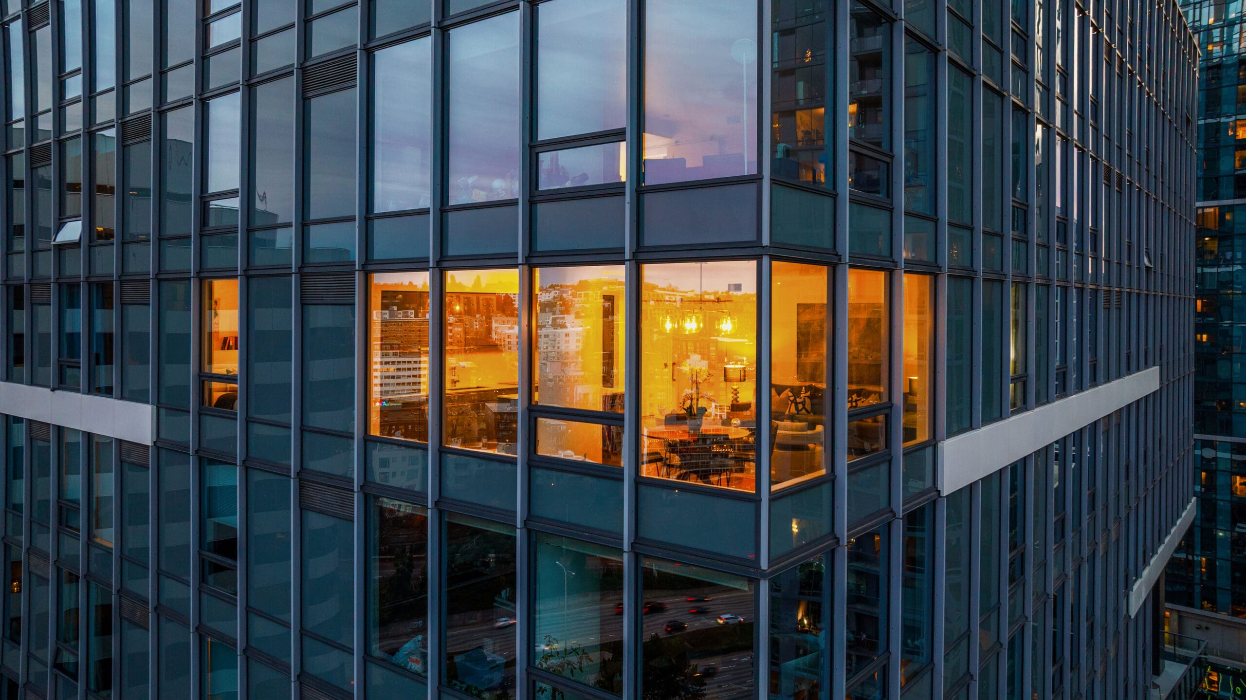 Modern urban exterior of a high-rise building in Seattle, showcasing large glass windows with warm interior lighting, reflecting a vibrant cityscape at twilight.