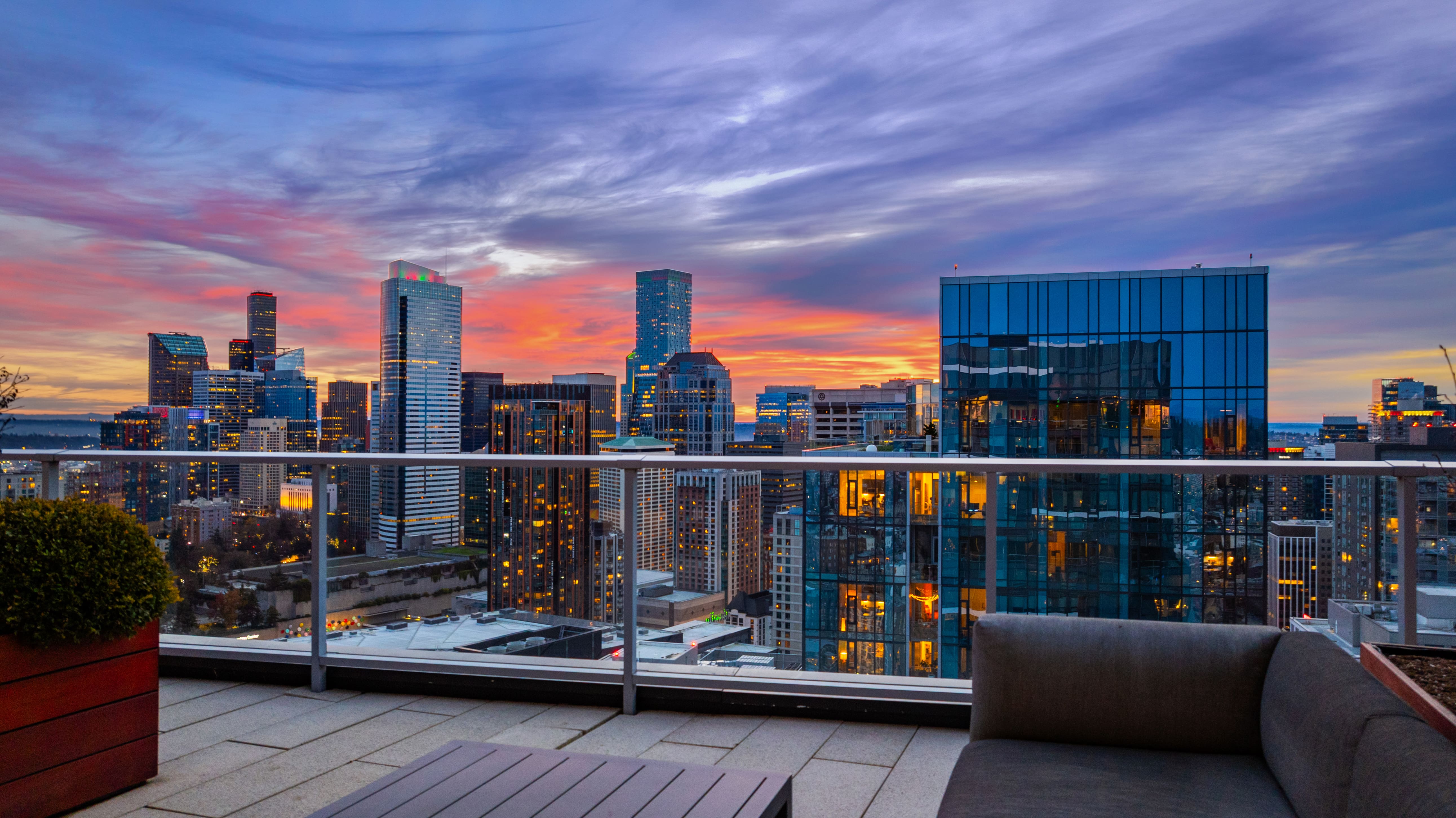 Rooftop terrace with modern seating and a stunning sunset view of Seattle's skyline, showcasing sleek glass buildings and vibrant colors in the Pacific Northwest sky.
