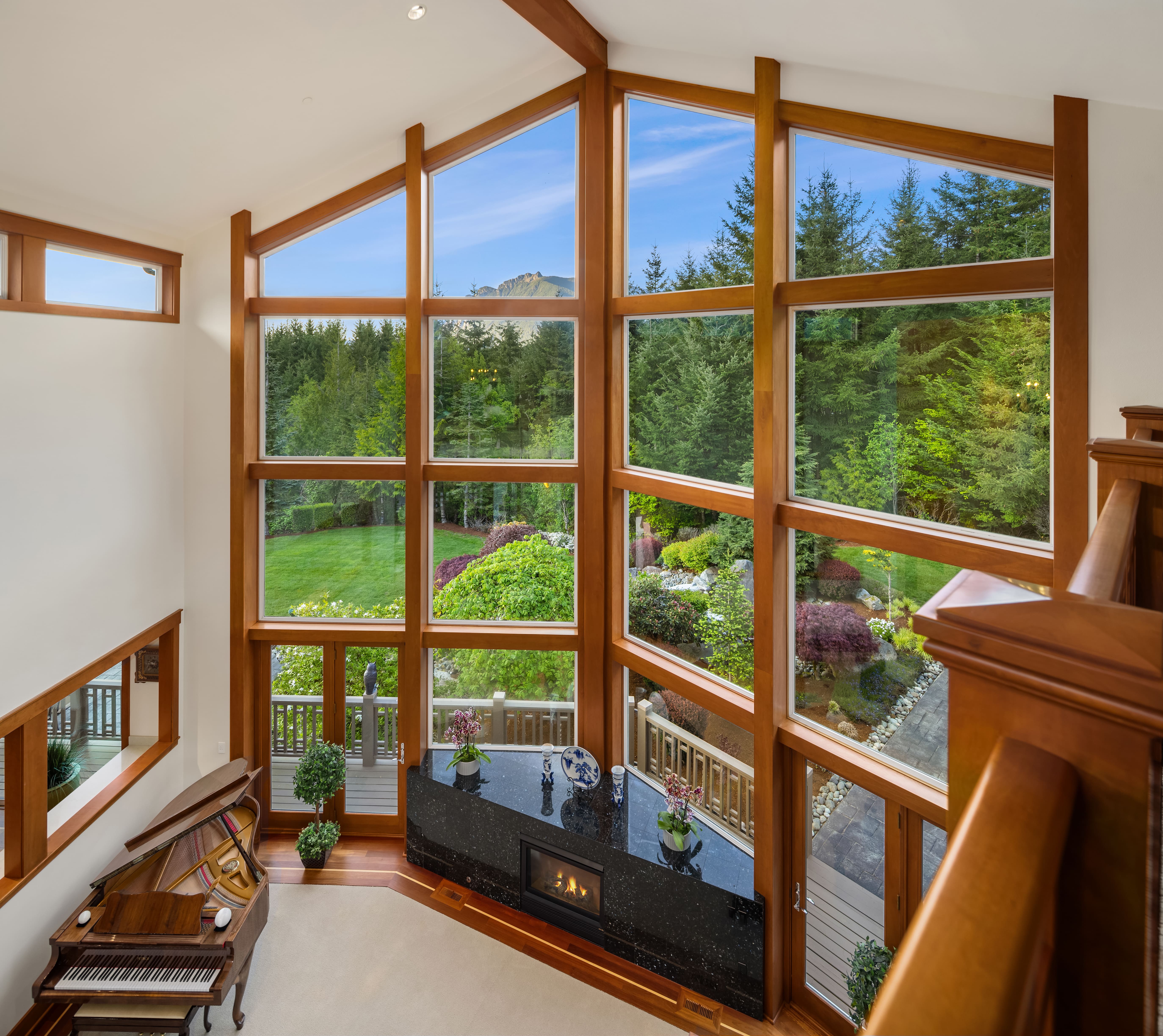 Elegant living room with large floor-to-ceiling windows showcasing lush greenery and landscaped gardens, featuring a grand piano and modern fireplace, located in the Pacific Northwest.