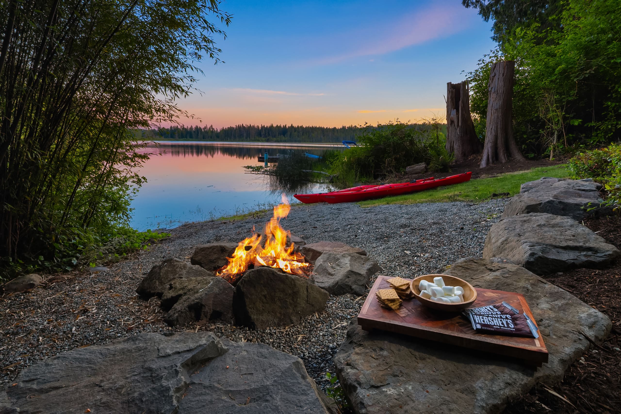 Cozy lakeside fire pit with s'mores ingredients, surrounded by natural greenery and a serene sunset view, located in the Pacific Northwest.