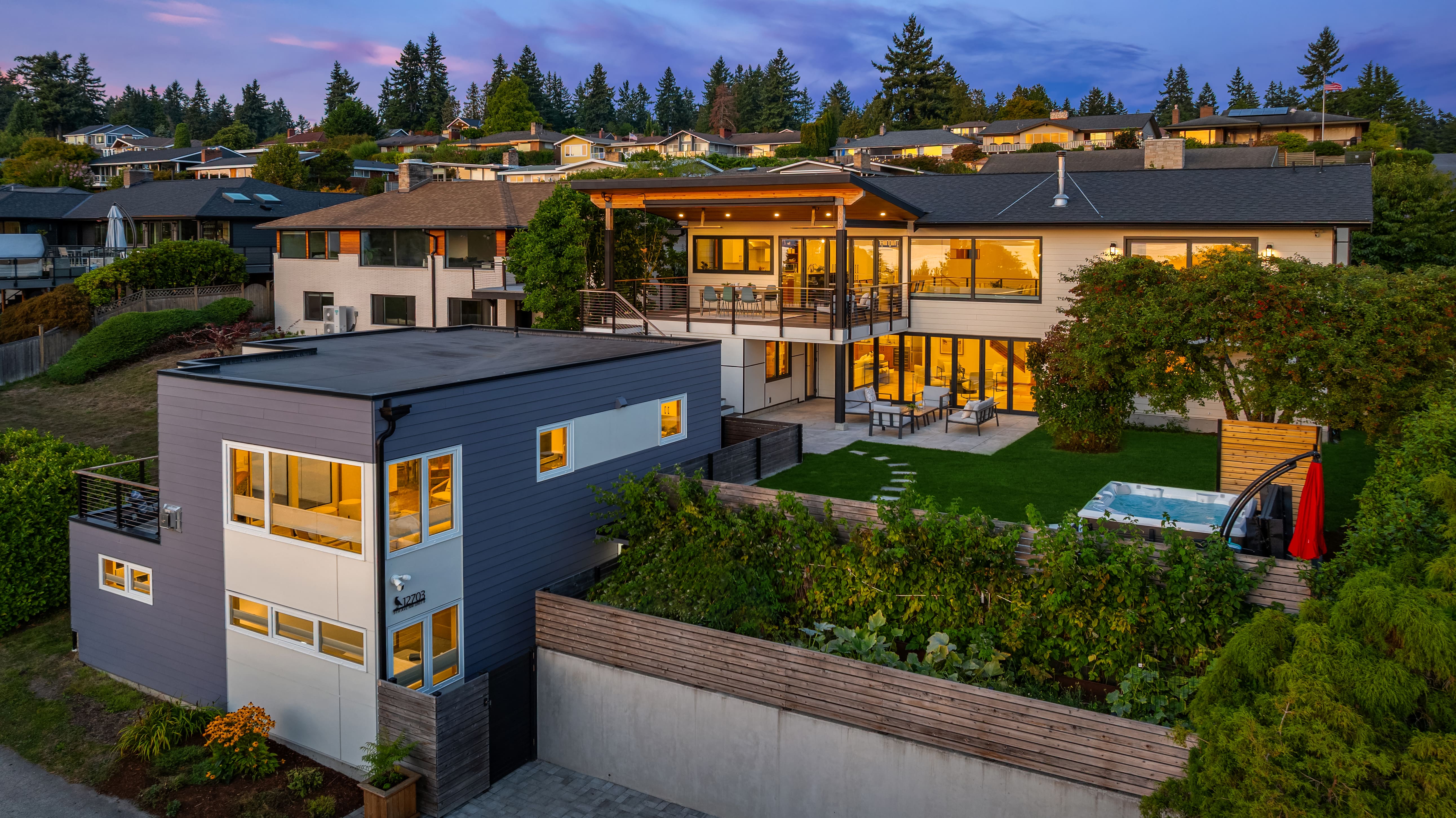 Modern exterior view of a luxurious home in the Pacific Northwest at twilight, featuring expansive outdoor living spaces, lush landscaping, and a hot tub, highlighting its elegant design and serene setting.